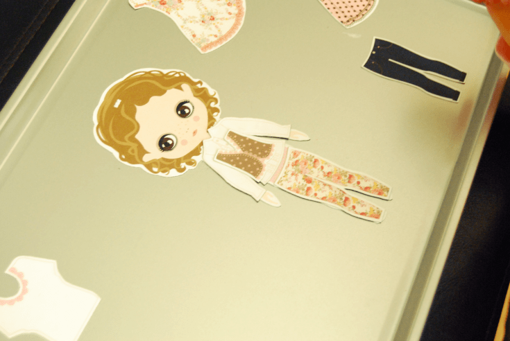 paper magnet doll with brown hair, a vest, long sleeve shirt and floral pants on a cookie sheet with a white shirt, a dress, and jeans.